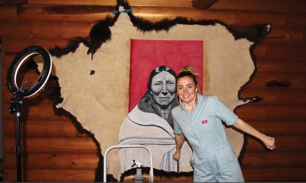 Artist Miah Chalfant, a light-skinned Native artist, smiles and poses in front of one of her tallow-hide paintings: a black-and-white portrait of a tribe matriarch in front of a deep, red square, all on a large, tan hide.