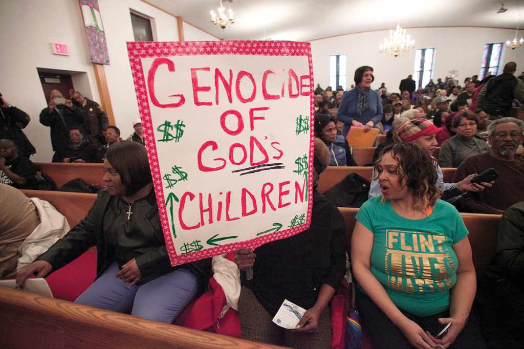 Residents of Flint, Michigan, gather at a prayer service inside a local church on Feb. 19, 2016. At the center of the frame, a congregant's face is obscured by a handmade sign reading "Genocide of God's children" in red font, with dollar signs surrounding the text. A woman to the right of the sign is wearing a green shirt with gold text reading "Flint Lives Matter."