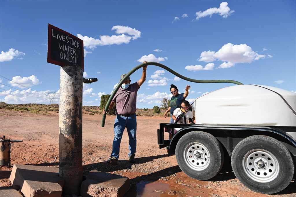 Navajo Nation resident Otto Tso (left), holds a long tube extending from a large portable water tank, helping Latoya Nez and her 6-year-old daughter, Arya Richardson, fill a water tank from a pump intended to provide water for livestock in Gap, Arizona. A sign on top of the pump reads "Livestock water only!!"