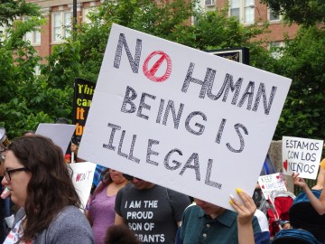 a person holds a square sign that reads No Human Being is Illegal at a protest