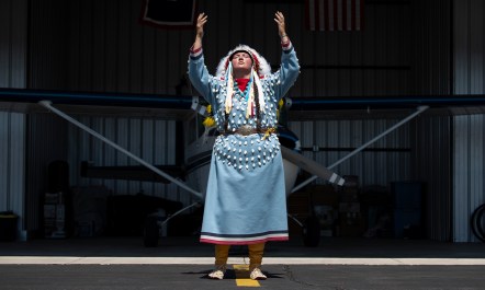 Shawnee Real Bird, in full regalia, poses in front of her airplane in a hangar. She is raising both her hands and looking skyward.