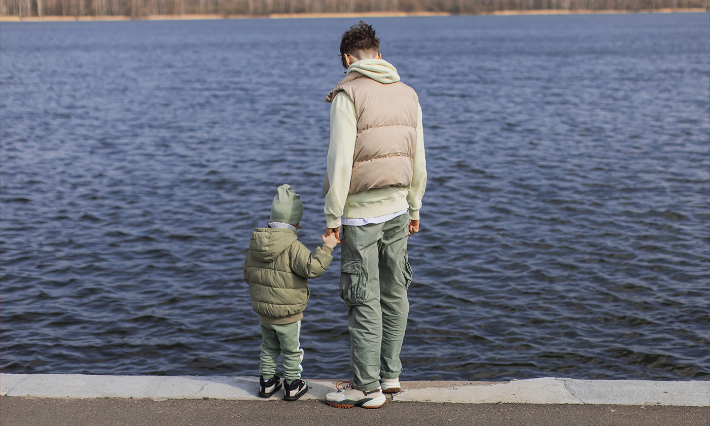 A photograph of an adult holding the hand of a small child looking out on a body of water.