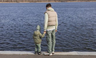 A photograph of an adult holding the hand of a small child looking out on a body of water.