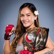Ilima-Lei Macfarlane, a Hawaiian woman with a flower in her hair, holds up a gloved fist with a championship belt over her shoulder