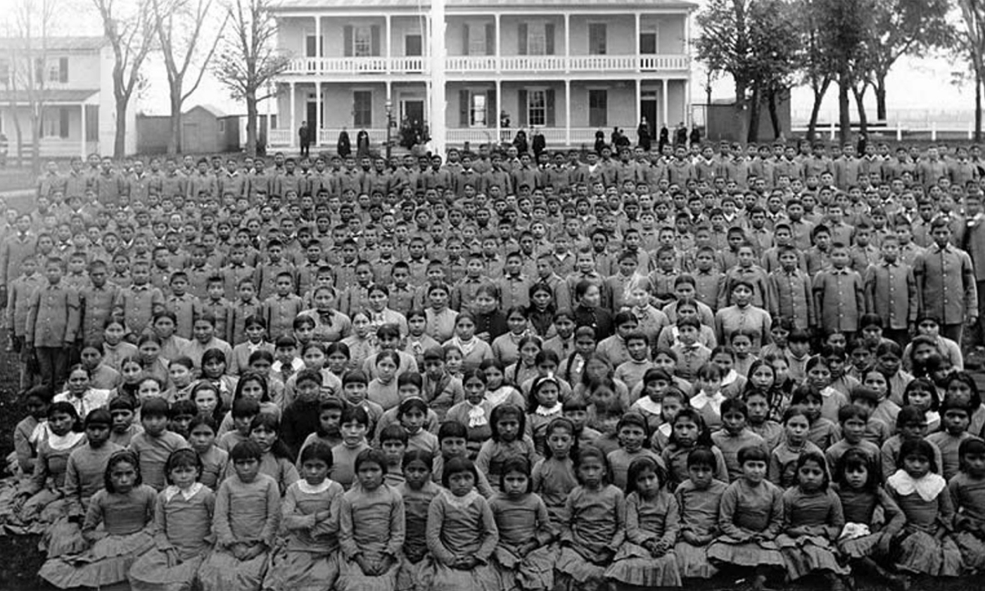 An undated group photo taken at Carlisle Indian Industrial School. At least one hundred Native children stand in front of large boarding school.