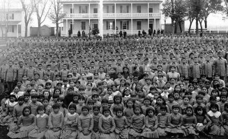 An undated group photo taken at Carlisle Indian Industrial School. At least one hundred Native children stand in front of large boarding school.