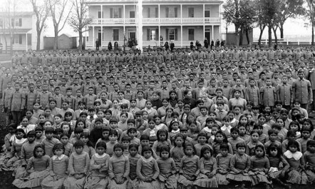 An undated group photo taken at Carlisle Indian Industrial School. At least one hundred Native children stand in front of large boarding school.