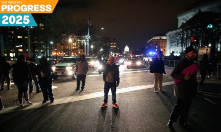 A night-time image of several protesters standing in a street as headlights in the distance illuminate them.
