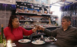 An image from the documentary Patrice: The Movie, which is available to stream on Hulu. Patrice, a black woman wearing a red shirt, has a toast with wine glasses with her partner Gerry, a white man in a grey sweater, both seated at a dinner.