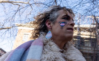 An older white person wearing trans pride earrings and facepaint stands outside on a sunny winter day. It is a picture from the Feb. 14, 2025 protest at the Stonewall Inn memorial.