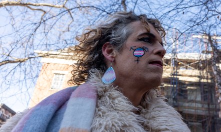 An older white person wearing trans pride earrings and facepaint stands outside on a sunny winter day. It is a picture from the Feb. 14, 2025 protest at the Stonewall Inn memorial.